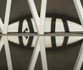 La simetría se rompe para dejar paso a lo esencial. Vista lejana de una madre y su hijo caminando entre columnas simétricas con reflejos en el agua.